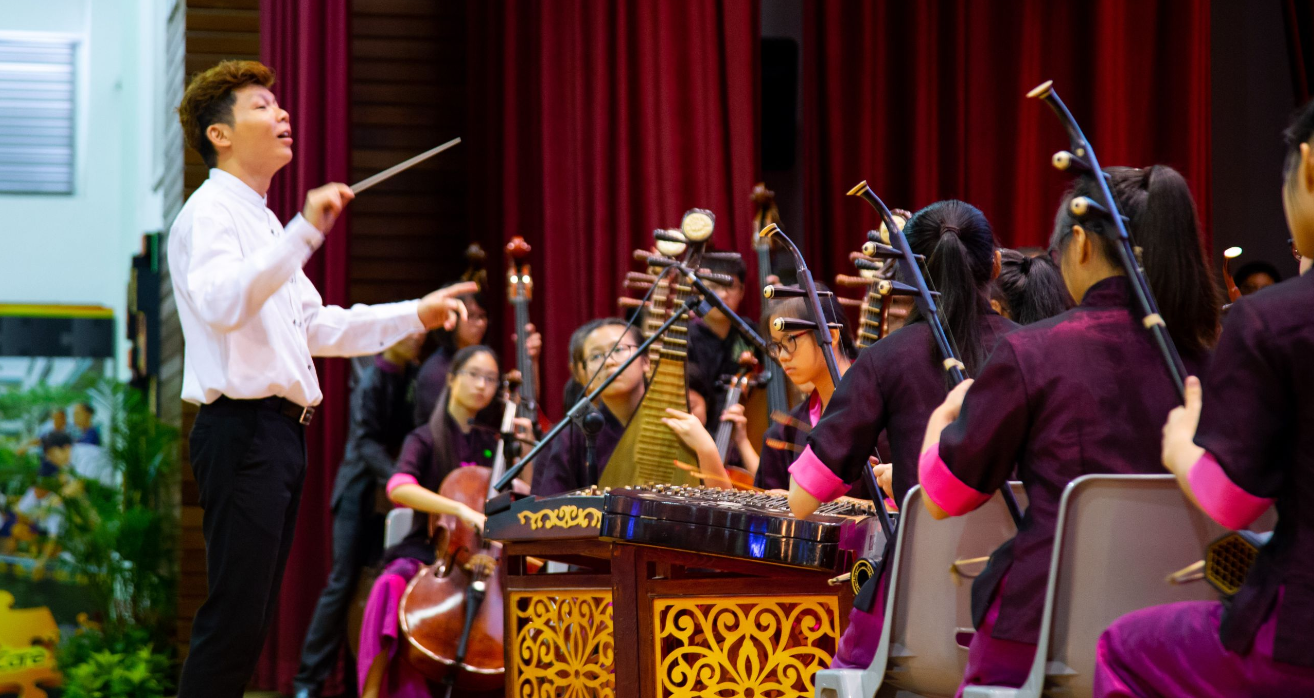 A conductor in a white shirt leads a youth Chinese orchestra playing traditional string instruments on stage.